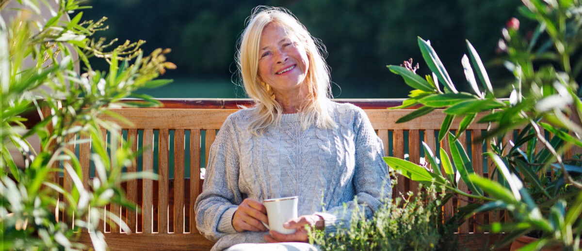 אישה מבוגרת על ספסל בשמש, בגינה, מחייכת - חוסן נפשי בגיל השלישי